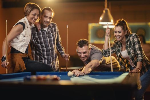 Young cheerful friends are playing billiards in bar after work. A couple that wins fame and the losers are in a bad mood.