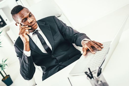 Pensive African businessman talking on smartphone and working on laptop in modern office.