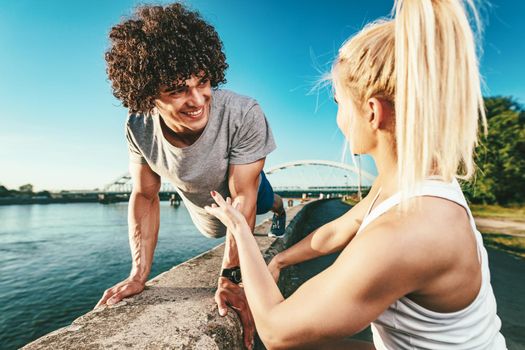 Young fitness couple is doing workout on the wall  by the river in a sunset. The man is crouching and holding kettlebell, and the woman supports him.