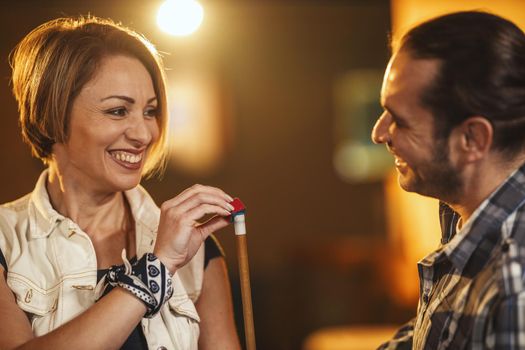 Smiling young woman apply chalk to the end of their cue sticks in billiard club.