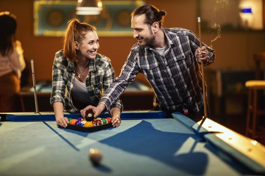 Young smiling cheerful couple is playing billiards in bar after work. They are having fun and involved in recreational activity.