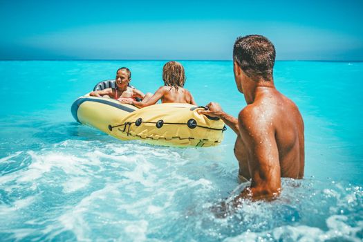 Happy family is enjoying floating in yellow kayak at tropical ocean water during summer vacation. The father is guarding children who are driving in a rubber boat. 
