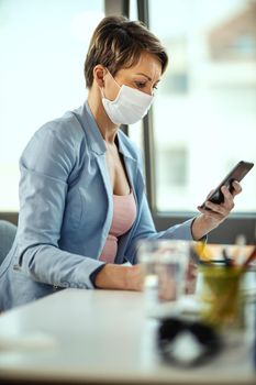 Business woman in a medical protective mask works from home at the computer, using smartphone, during self-isolation and quarantine to avoid infection during flu virus outbreak and coronavirus epidemic.