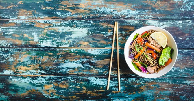 Asian noodles with vegetables, bowl, rustic wooden old background. Space for text. Top view. Soba noodles, vegetables, mushrooms, chopsticks. Vegetarian/Vegan noodles. Asian style dinner with noodles