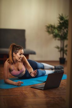 Young smiling woman is records a internet tutorial and doing sports training in the living room on floor mat in front of the laptop.