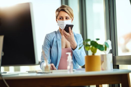 Business woman in a medical protective mask works at the computer and talking on a smartphone during self-isolation and quarantine to avoid infection during flu virus outbreak and coronavirus epidemic.