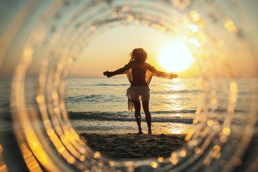 A beautiful creative composition of a sea landscape shot through a circle focus showing a young woman who is having fun on the beach in sunset.