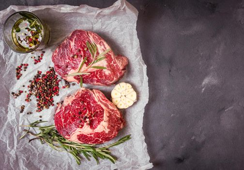 Raw juicy ribeye steaks with seasonings ready for roasting on baking paper. Rustic concrete background. Fresh marbled meat steaks with herbs, garlic, olive oil, pepper, salt. Space for text. Top view