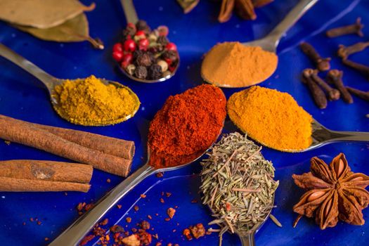 Different colorful spices in little spoons on dark blue background. Curry, turmeric, cinnamon sticks, bay leaves, sumac, cloves, peppercorn, paprika, rosemary, star anise. Top view. Closeup