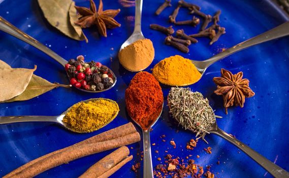 Different colorful spices in little spoons on dark blue background. Curry, turmeric, cinnamon sticks, bay leaves, sumac, cloves, peppercorn, paprika, rosemary, star anise. Top view. Closeup