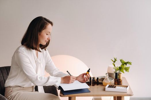 The aromatherapist girl is sitting in her office and holding a bottle of aromatic oil in her hands and writing something down. there are essential oils on the table.