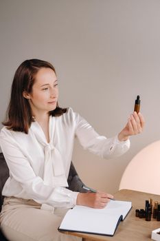 The aromatherapist girl is sitting in her office and holding a bottle of aromatic oil in her hands and writing something down. there are essential oils on the table.