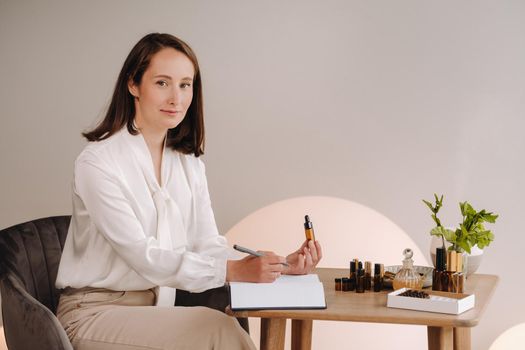 The aromatherapist girl is sitting in her office and holding a bottle of aromatic oil in her hands and writing something down. there are essential oils on the table.