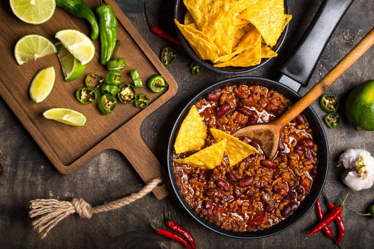 Chili con carne in frying pan on dark wooden background. Ingredients for making Chili con carne. Top view. Chili with meat, nachos, lime, hot pepper. Mexican/Texas traditional dish Chili con carne