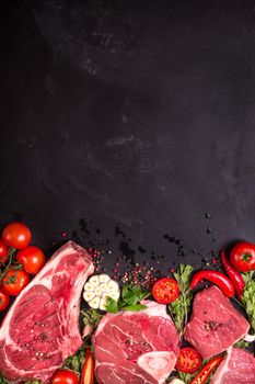 Raw juicy meat steaks ready for roasting on a black chalk board background. Rib eye steak on the bone, veal shank (ossobuco), fillet with cherry tomatoes, hot pepper and herbs. Space for text