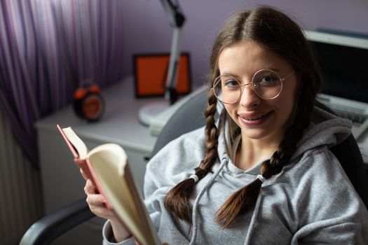A cheerful girl is happily reading her favorite book. The brunette has her glasses on and her hair is styled in two braids. She loves reading books.