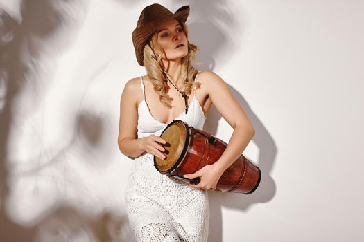 Beautiful Blonde Young Woman Wearing White Crochet Dress and Bown Cowboy Hat under the Shade of Foliage. Bohemic Country Style. Free People in Boho Outfit. Music Gypsy Woman Dancing with Drum