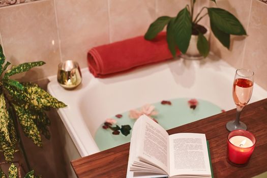 Time for Yourself. Relax at home. Bath tub with flower petals. Book, candles and glass of wine on a wood tray. Organic Spa Relaxation in comfort cozy bathroom