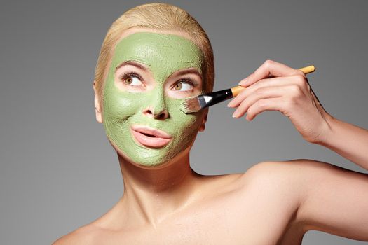 Beautiful Funny Woman Applying Green Facial Mask. Beauty Treatments. Close-up Portrait of Spa Girl Apply Clay Facial mask on grey background. Emotional Female Face with Cute Surprised Grimace