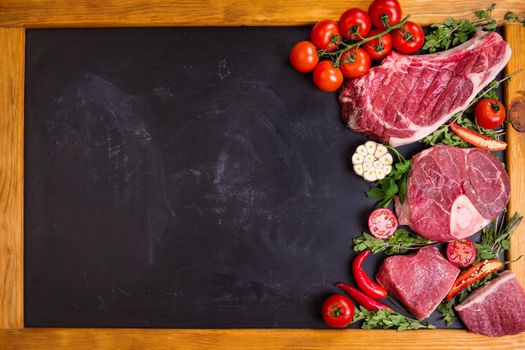 Raw juicy meat steaks on a black chalk board background with wooden frame. Rib eye steak on the bone, veal shank (ossobuco), fillet with cherry tomatoes, hot pepper and herbs. Space for text