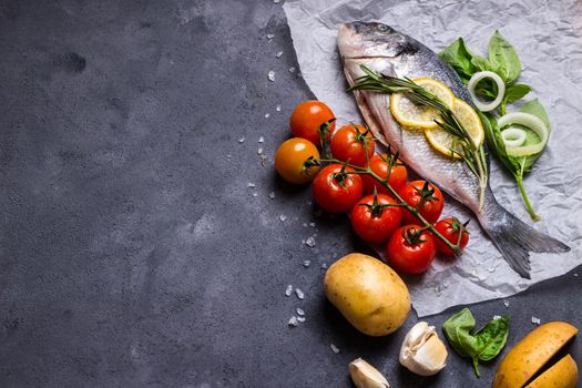 Raw fish with fresh ingredients ready to cook. Fish, lemon, herbs, potato, tomatoes. Ingredients for cooking on dark rustic background. Space for text. Diet and healthy food. Fish background. Top view