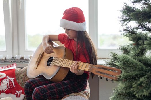 Happy little girl in red hat playing the on the background of christmas tree. Talent kid having fun on winter holidays at cozy decorated home