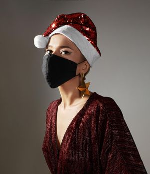 Woman with red christmas hat, black medical mask. Fashion style with shiny clothes and accessories for xmas time. Protect yourself from Coronavirus even on holidays. Merry Christmas and Happy New Year