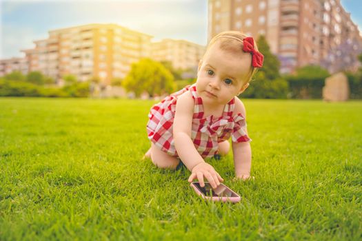 Curious little baby boy with blond curly hair dressed in light summer clothes sitting on lawn with fresh green grass sunny day playing on mobile phone, horizontal picture. High quality photo