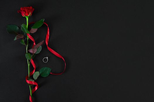 One dark red rose with red ribbon and the ring on black background. Romantic Valentines holidays concept. Valentine's day greeting card. Top view. Copy space. Still life. Flat lay