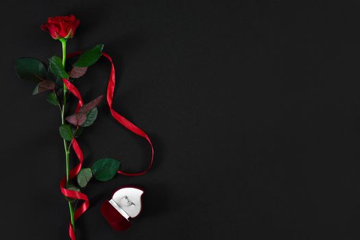 One dark red rose with red ribbon and box with ring on black background. Romantic Valentines holidays concept. Valentine's day greeting card. Top view. Copy space. Still life. Flat lay