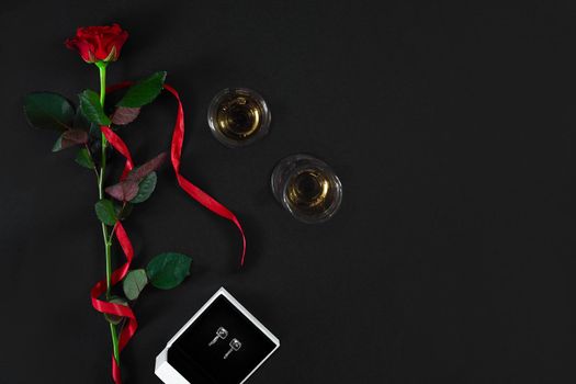 One dark red rose with red ribbon and box with earrings on black background. Romantic Valentines holidays concept. Valentine's day greeting card. Top view. Copy space. Still life. Flat lay