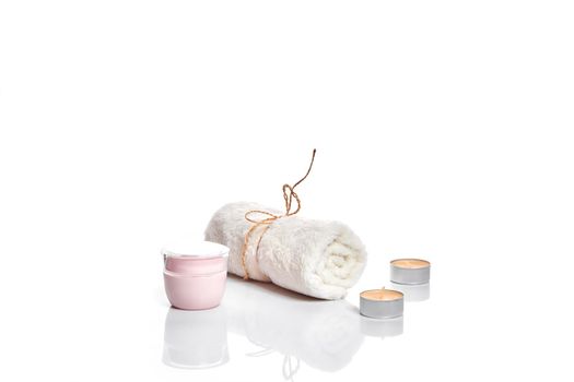 Glass jar with face cream, pink color. White towel and candles on light background. Still life. Copy space.