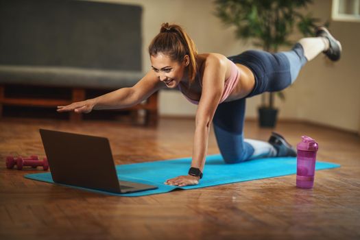 Young smiling woman is doing fitness exercises in the living room on floor mat at home, looking at the laptop, in morning sunshine.