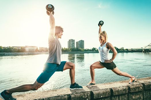 Young fitness couple is doing workout with dumbbells  on the wall  by the river in a sunset.