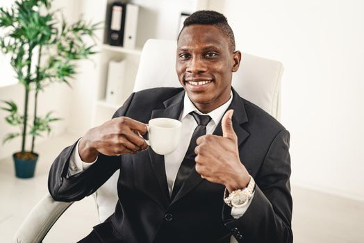 African businessman is sitting in the office and drinking coffee. He is satisfied with success and show thumb up.