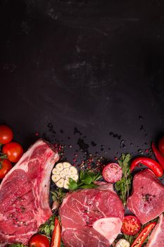 Raw juicy meat steaks ready for roasting on a black chalk board background. Rib eye steak on the bone, veal shank (ossobuco), fillet with cherry tomatoes, hot pepper and herbs. Space for text