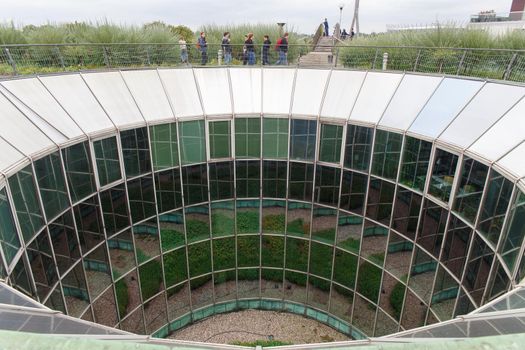 Warsaw, Poland - July 16, 2019: Tourists walking on roof fully glazed rounded University of Warsaw Library. Numerous reflections of the courtyard with landscaping create a whimsical pattern.