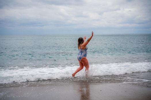 A plump woman in a bathing suit enters the water during the surf. Alone on the beach, Gray sky in the clouds, swimming in winter