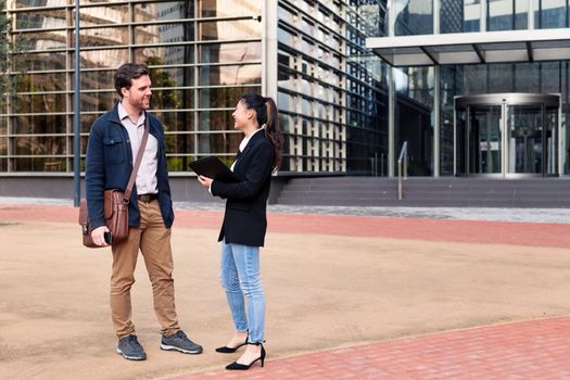caucasian man and asiatic woman smiling and talking about business in front of an office building, concept of coworkers and lifestyle, copy space for text