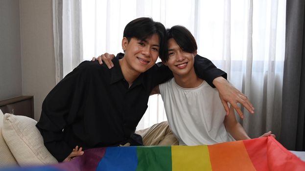 Cheerful homosexual couple embracing and holding LGBT pride flag with love together. LGBT, pride, relationships and equality concept.