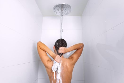 Rear view of young woman taking shower and washing hair with shampoo under water falling from rain shower head. High quality photo