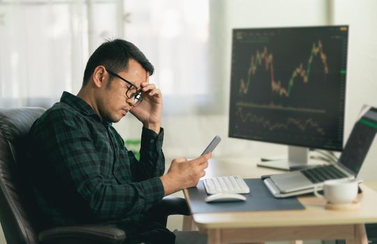 Asian man trader unhappy and serious sitting at home office in front of monitors with cryptocurrency graph holding smartphone monitoring cryptocurrency price