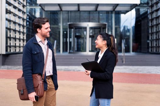 happy caucasian man and smiling asian woman talking about business in front of the entrance to an office building, concept of coworkers and lifestyle