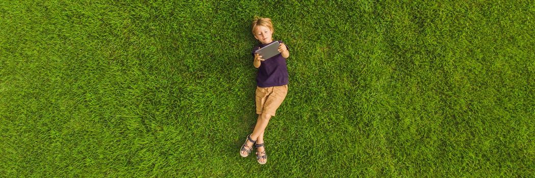 Boy lying on the grass in the park and looks at the tablet. Photos from the drone, quadracopter. BANNER, LONG FORMAT