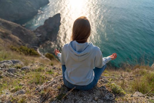 Woman tourist enjoying the sunset over the sea mountain landscape. Sits outdoors on a rock above the sea. She is wearing jeans and a blue hoodie. Healthy lifestyle, harmony and meditation.