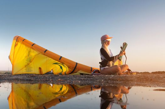 Attractive stylish young caucasian woman in cap sunglasses and kitesurfer outfit sits on the sandy shore next to her kite at sunset reflecting in the water.