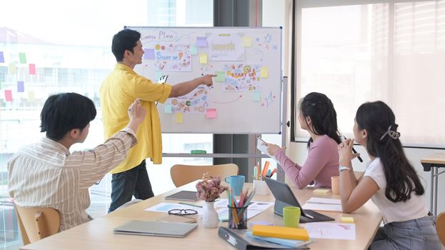 Asian male manager presenting business plan information on white board to diverse employees at office meeting.