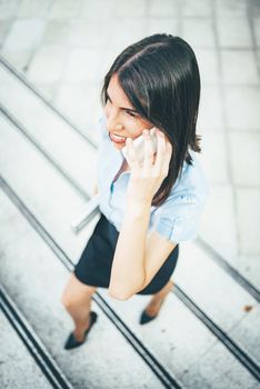 Young beautiful businesswoman walking up the stairs in front of office building, holding a laptop and talking on smartphone.