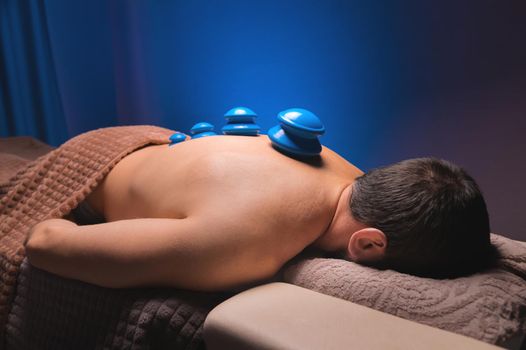 Close-up of a man's back with rubber vacuum cans installed in a dark room of a spa salon.