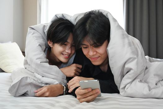 Happy young gay couple lying under blanket on bed and using mobile phone together. LGBT and love concept.